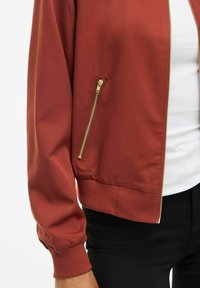 Leicht rostfarbener Jacke mit goldenem Reißverschluss-Taschen und Manschetten-Details, getragen über einem weißen Hemd und schwarzen Hosen, gezeigt von der Taille bis zum Ellebogen.