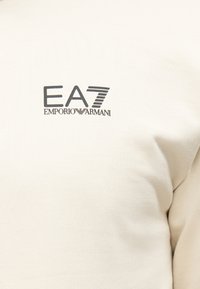 Ύφασμα κρεμ με μαύρο λογότυπο "EA7 Emporio Armani" τυπωμένο στην περιοχή του άνω στέρνου ενός ενδύματος.