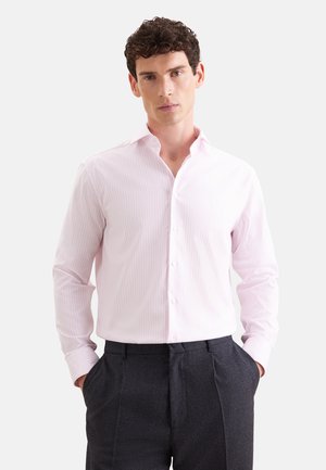 Man met krullend donker haar die een lichtroze gestreept overhemd met knopen en donkergrijze broek draagt, staand met handen in de zakken.