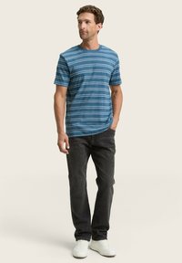 Camiseta azul a rayas de mangas cortas, hecha de algodón. Combinada con jeans oscuros y zapatillas blancas, de pie frente a un fondo claro.
