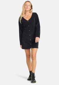 Marineblaues Cordkleid mit langen Ärmeln, V-Ausschnitt, vorderer Knopfleiste und zwei Seitentaschen, kombiniert mit schwarzen Kampfstiefeln.