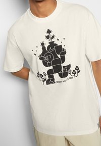 Krémové bavlněné tričko s černým grafickým designem stylizovaných postav, květin a textu "Bloom and ZOOM." Krátké rukávy.