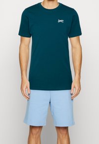 Tyrkysové bavlněné tričko s krátkým rukávem, se stříbrnou grafikou nůžek na levé straně hrudi. Vytvořeno s lehkými modrými šortkami.