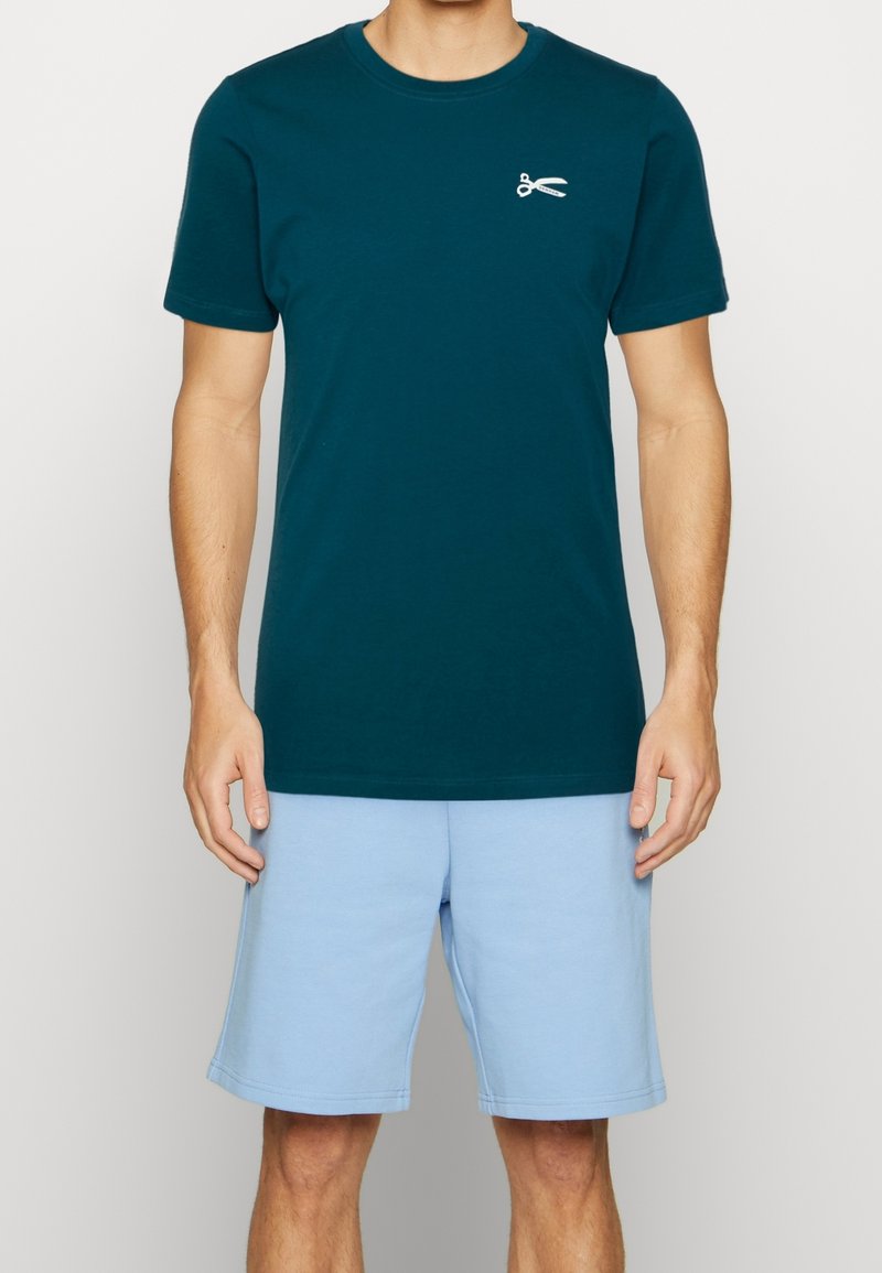 Tyrkysové bavlněné tričko s krátkým rukávem, se stříbrnou grafikou nůžek na levé straně hrudi. Vytvořeno s lehkými modrými šortkami.