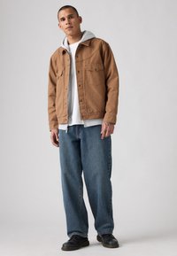 Brun denimjakke med grå hættetrøje underneath, hvid t-shirt og blå loose-fit jeans. Sorte sko fuldender outfittet.
