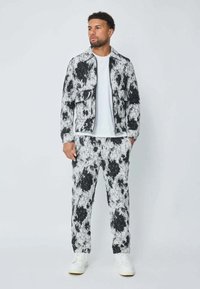Homme debout portant une veste et un pantalon à motifs noir et blanc avec une chemise blanche et des baskets blanches sur fond uni.