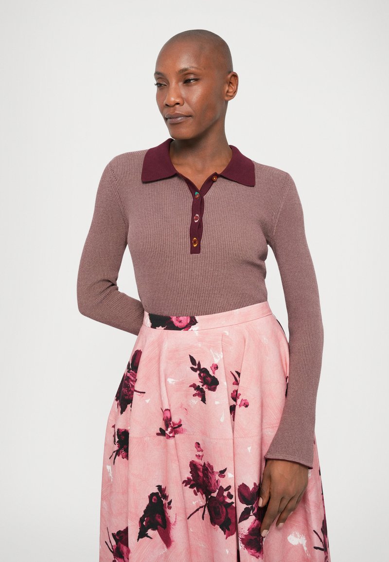 Långärmad ribbad topp i mauve med en bordeauxfärgad krage och detaljer med knappar, tillsammans med en rosa blommig plisserad kjol med mörka accenter.