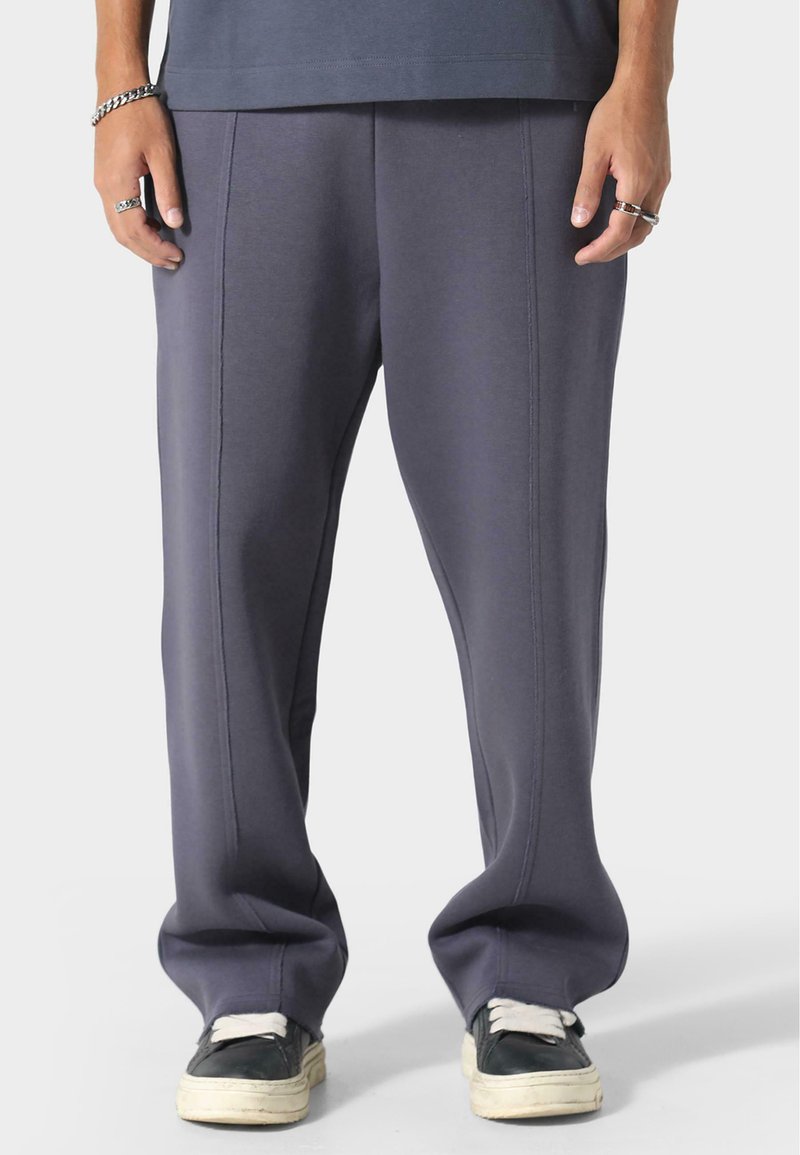 Pantalon de survêtement gris à coupe décontractée, fabriqué en mélange de coton doux, avec des coutures latérales et un design de jambe fuselée. Associé à des baskets noires.