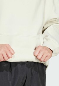 Felpa color crema con polsini e orlo a costine, caratterizzata da un tessuto texturizzato. Mani che afferrano l'orlo, abbinata a pantaloni scuri e con motivi.