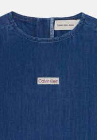 Džinsa T-krekls tumši zilā krāsā ar apaļu kakla izgriezumu un Calvin Klein logotipa uzliku baltā un sarkanā krāsā, ar redzamām šūšanas detaļām.