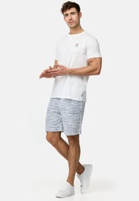 Camiseta blanca de manga corta con un pequeño logo, combinada con pantalones cortos azules estampados con diseños geométricos, completada con zapatillas blancas.