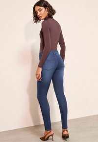 Bodysuit de manga larga marrón combinado con jeans skinny azules. Los jeans tienen bolsillos traseros y una cintura alta; el bodysuit tiene un diseño ajustado.