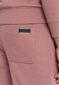 Rosa Jogginghosen aus weichem Material, mit einer Rückentasche und einem schwarzen Etikett. Das Design umfasst gerippte Bündchen und Säume.