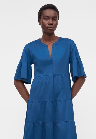 Γυναίκα με κοντά σγουρά μαλλιά που φοράει μπλε φόρεμα με βολάν μέχρι τον αγκώνα, στέκεται μπροστά σε απλό φόντο.