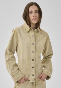 Frau mit braunen Haaren trägt beige Jeansjacke und passende Hose, steht mit den Händen in den Taschen vor einem hellgrauen Hintergrund.