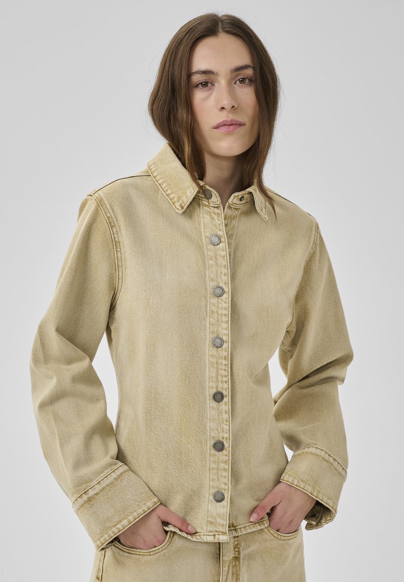 Frau mit braunen Haaren trägt beige Jeansjacke und passende Hose, steht mit den Händen in den Taschen vor einem hellgrauen Hintergrund.