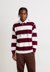 Fiatal férfi fonott hajú, hosszú ujjú rúgby pólót visel, széles bordó és fehér vízszintes csíkokkal, barna nadrággal a fehér háttér előtt.