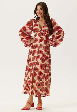 Kvinde smilende, iført en lang blomsterkjole med voluminøse ærmer og røde sandaler, stående med hænderne på hofterne mod en ensfarvet baggrund.