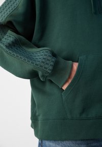Grønn hettegenser med teksturerte ermeakkenter, ribbestrikkede mansjetter og en frontlomme. Laget av mykt stoff, designet for komfort og varme.
