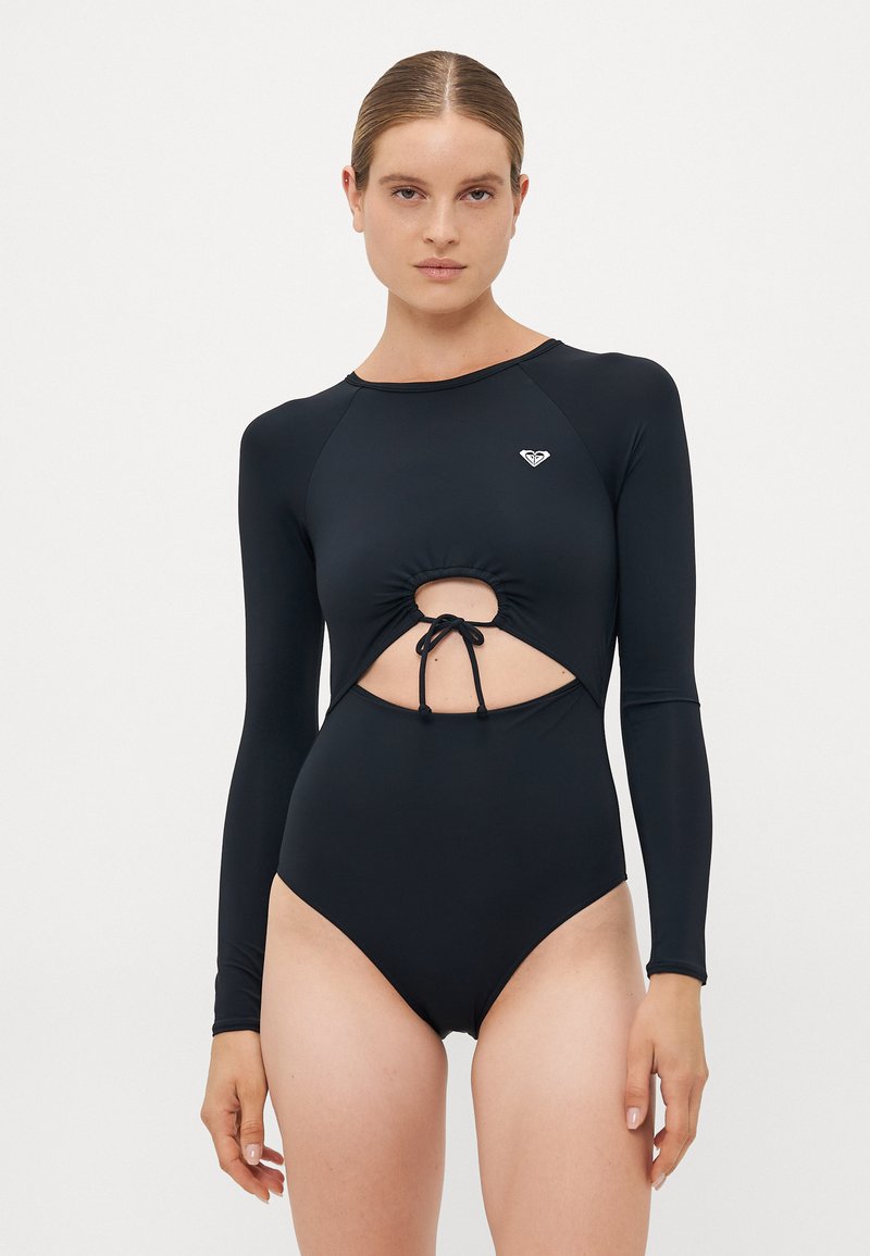 Mulher vestindo um fato de banho preto de mangas longas com um recorte frontal e detalhe de laço, de pé contra um fundo branco liso.