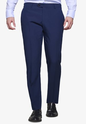 Mężczyzna ubrany w granatowe, dopasowane eleganckie spodnie, jasnoniebieską koszulę na guziki oraz czarne skórzane buty, stojący na białym tle.