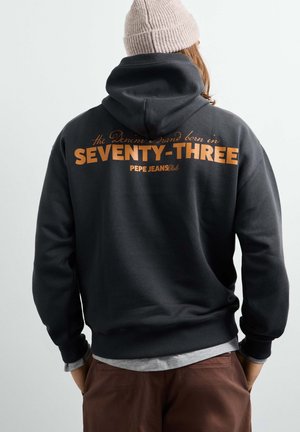 Persoon draagt een zwarte hoodie met oranje tekst "SEVENTY-THREE" en "PEPE JEANS club" op de rug en een beige gebreide muts.