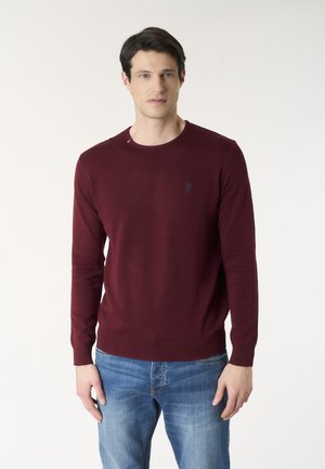 Uomo che indossa un maglione bordeaux a maniche lunghe e jeans blu, in piedi davanti a uno sfondo chiaro uniforme, rivolto in avanti.
