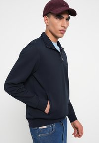 Marine blauwe rits-hoodie met lange mouwen en zijzakken, gedragen met een bordeauxrode pet. Heeft een eenvoudig ontwerp en een casual pasvorm.