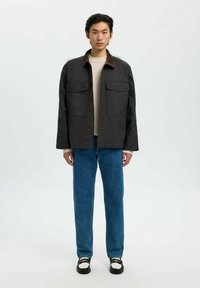Mörkbrun, lätt jacka med två bröstfickor, kombinerad med en krämfärgad tröja och blå raka jeans; svartvita skor.