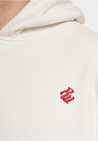Felpa con cappuccio crema realizzata in un morbido tessuto, con un logo ricamato rosso sul petto, cappuccio con cordino e dettagli di cucitura sottili.