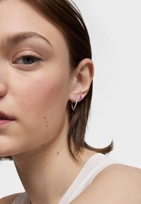 Mujer joven con cabello corto y castaño, que lleva pequeños pendientes de aro plateados y de piedra rosa, mostrada en primer plano sobre un fondo liso.