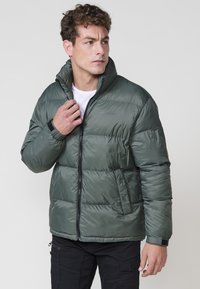 Chaqueta acolchada verde tipo puffer con alto cuello, cierre frontal y puños elásticos. Cuenta con bolsillos laterales y un logo sutil en el pecho.