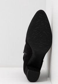 Bottine noire en daim avec semelle en caoutchouc à motif et petit talon carré, positionnée verticalement contre des murs blancs d'angle.