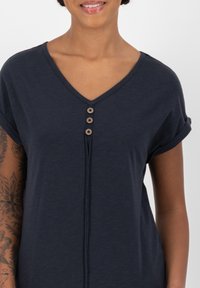 T-shirt blu navy a V in morbido tessuto, con maniche arrotolate e quattro bottoni decorativi lungo la pattina frontale.