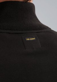 Schwarzer Rollkragenpullover mit gerippter Textur, ausgestattet mit einem rechteckigen Etikett mit "PME LEGEND" in gelber und schwarzer Stickerei auf der Rückseite.