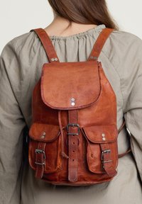 Hnědý kožený batoh s klopou, dvěma předními kapsami, kovovými stříbrnými akcenty a nastavitelnými polstrovanými popruhy. Texturovaný povrch.