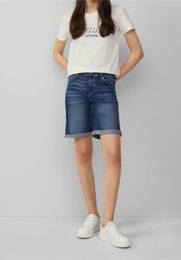 Denim shorts, rolled cuffs, dark blue color, paired with a light beige t-shirt featuring a printed design, and white sneakers.