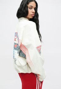Převelký krémový svetr s barevnými grafikami a červenými pruhovanými akcenty. Obsahuje trojúhelníkovou nášivku a volný střih.