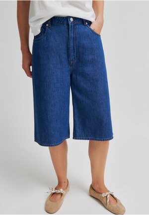 Person som har på seg knelang blå denimshorts, en hvit skjorte stukket inn og beige ballettsko med bånd.