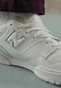 Bílé tenisky s hladkým koženým svrškem, perforovanými detaily a reflexním logem "N". Obsahují bílé tkaničky a texturovanou gumovou podrážku.