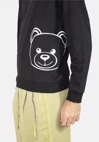 Černá mikina s bílým potiskem medvědí hlavy na boku, s žebrovaným výstřihem a manžetami. Kombinováno se světle zelenými kalhotami se zipy.