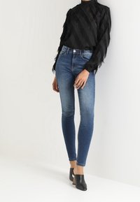 Top negro de manga larga con rayas texturizadas, combinado con jeans de mezclilla azul ajustados. La modelo lleva puestos tacones negros de punta abierta con un diseño simple.