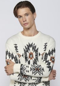 Kreemist pullover, millel on geomeetrilised mustad ja pruunid mustrid, soonikud vöökohtades ning ümmargune kaelus, valmistatud pehmest kudumaterjalist.