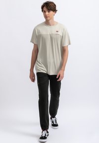 Lichtbeige T-shirt met korte mouwen en een kleine grafische print op de borst, gecombineerd met donkergroene tapered joggers en zwarte sneakers.