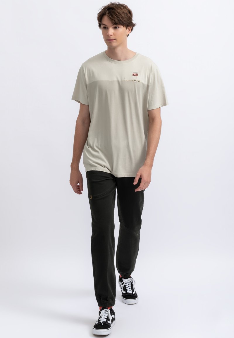 Lichtbeige T-shirt met korte mouwen en een kleine grafische print op de borst, gecombineerd met donkergroene tapered joggers en zwarte sneakers.