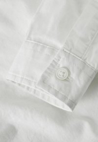 Witte katoenen shirtmanchet met een gladde textuur, voorzien van een enkele witte knoop en nette stiksels langs de rand.