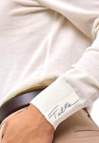 Kremowy top z długim rękawem i prążkowanymi mankietami. Mankiety ozdobione naszywką z napisem „Falke” w kolorze szarym. Gładka, lekka tkanina.