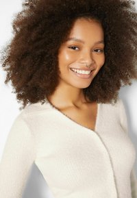 Vit fluffig kofta med v-ringning, texturerad tyg, sju knappar fram och subtila ribbade detaljer runt axlarna.