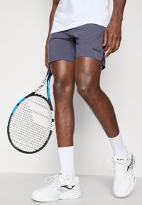 Hallitushedastatud viimistlusega hallid spordialused lühikesed püksid ja logo, koos valgete tennisekingade ja lühikeste sokkidega. Vasakus käes on tennisereket.