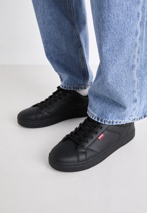 Czarne skórzane trampki Levi's noszone z jasnoniebieskimi dżinsami na jasnej podłodze.